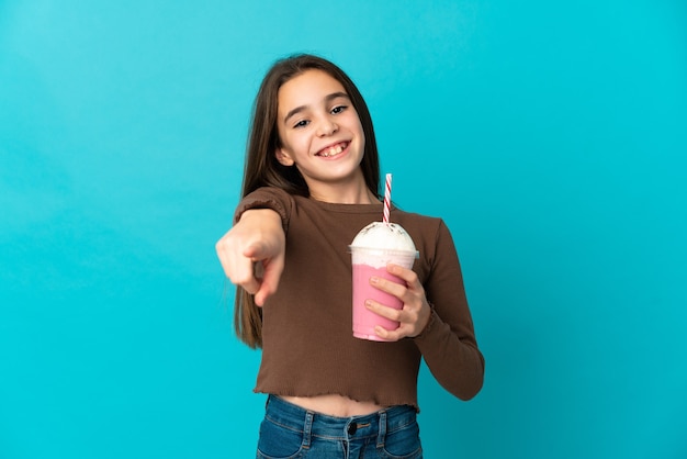 Little girl with strawberry milkshake isolated on blue wall pointing front with happy expression