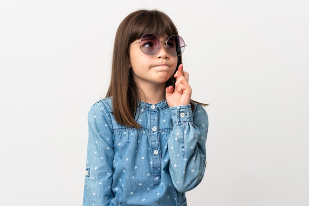 Little girl with sunglasses isolated on white background with fingers crossing and wishing the best