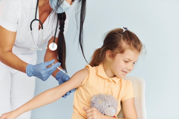 Фото Маленькая девочка с плюшевым мишкой врач в форме делает пациенту вакцинацию
