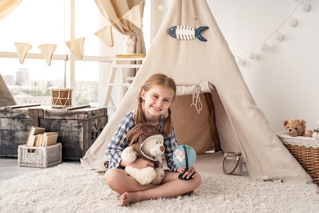 Little girl with teddy bear and globe sitting in wigwam settled in playroom