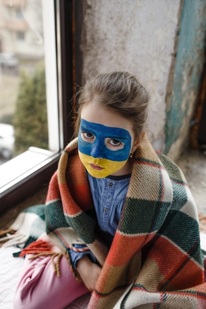 A little girl with a Ukrainian flag painted on her face is sitting covered with a blanket