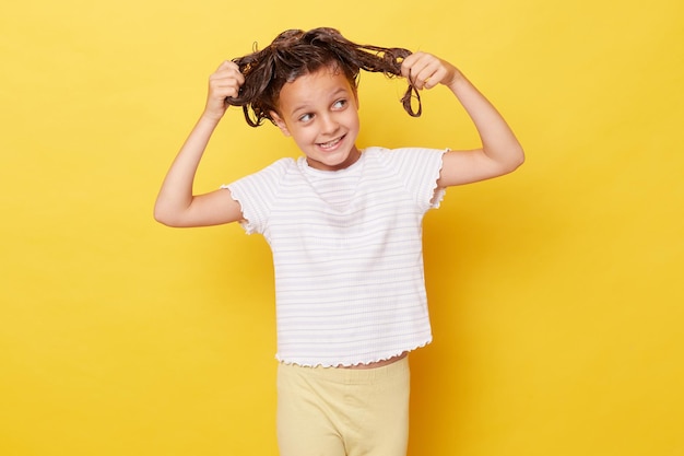 Little girl with wet hair wearing casual white Tshirt standing isolated over yellow background tangled hair while washing looking away with confused smile