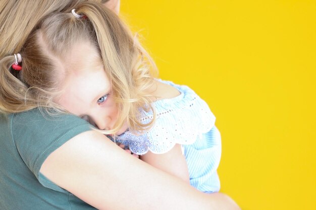 A little girl with white hair is upset and sad on her mother's shoulder on a yellow background Children's fears resentments motherchild relationships Mother and child motherhood