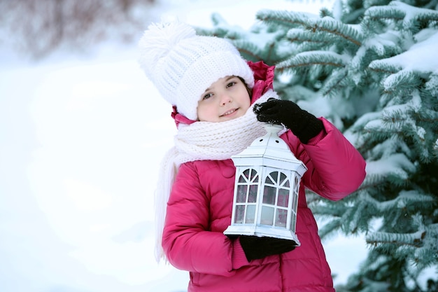 Little girl with winter clothes holding white lantern near fir tree in snowy park outdoor