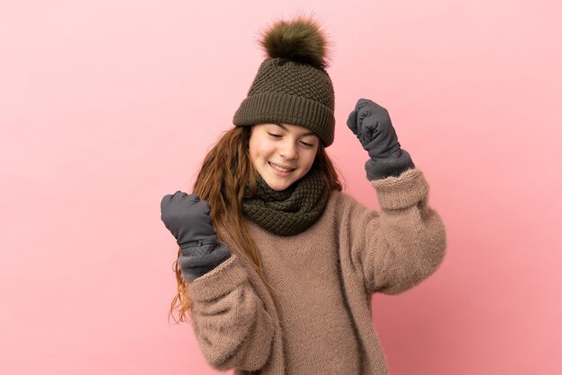 Little girl with winter hat isolated on pink background celebrating a victory