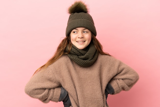 Bambina con cappello invernale isolato su sfondo rosa in posa con le braccia all'anca e sorridente