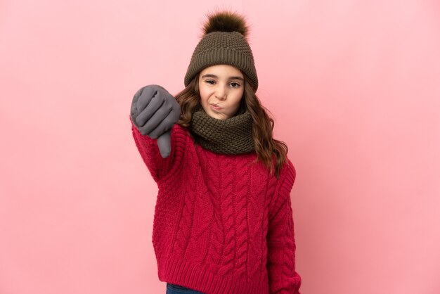 Little girl with winter hat isolated on pink background showing thumb down with negative expression