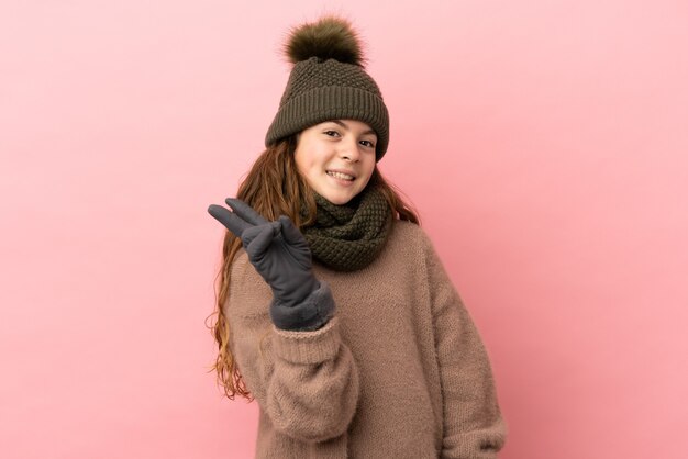 분홍색 배경에 격리된 겨울 모자를 쓴 어린 소녀가 웃고 승리 기호를 보여줍니다.