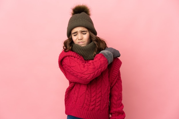 Маленькая девочка в зимней шапке изолирована на розовой стене и страдает от боли в плече из-за того, что приложила усилие