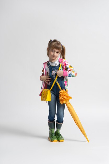 A little girl with a yellow handbag in casual autumn clothes and rubber boots plays with a folded umbrella and poses on a white background