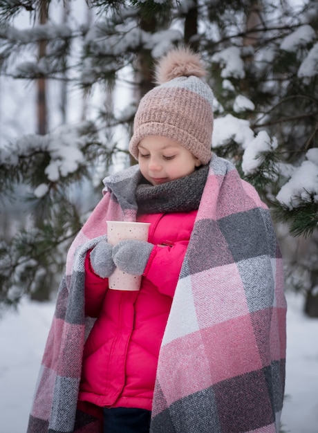 Little girl wrapped in a blanket drinking tea in the winter forest