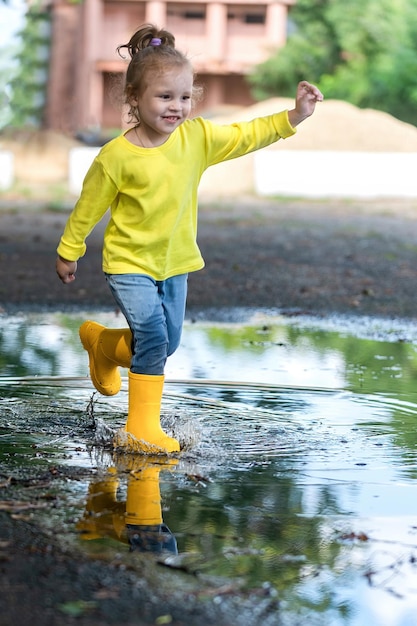 a little girl in yellow clothes and rubber boots runs merrily through the puddles after the rain