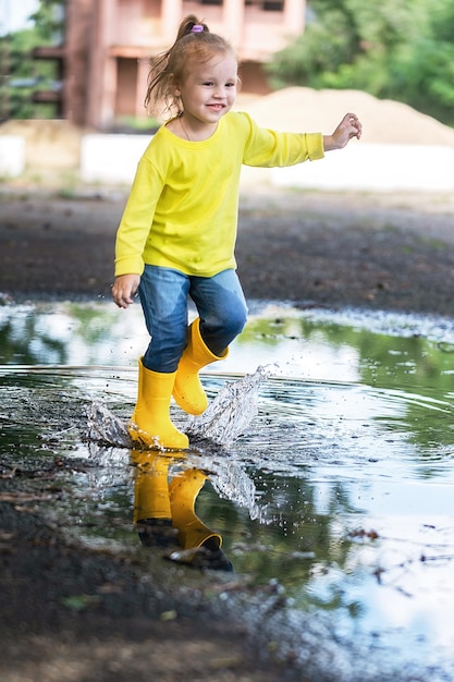 노란 옷과 고무 장화를 입은 어린 소녀가 비가 온 후 웅덩이를 즐겁게 뛰어다닌다