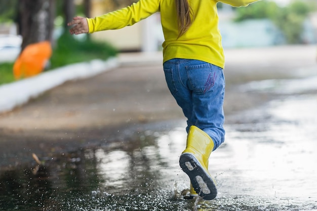 A little girl in yellow clothes and rubber boots runs merrily\
through the puddles after the rain