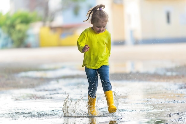 A little girl in yellow clothes and rubber boots runs merrily\
through the puddles after the rain