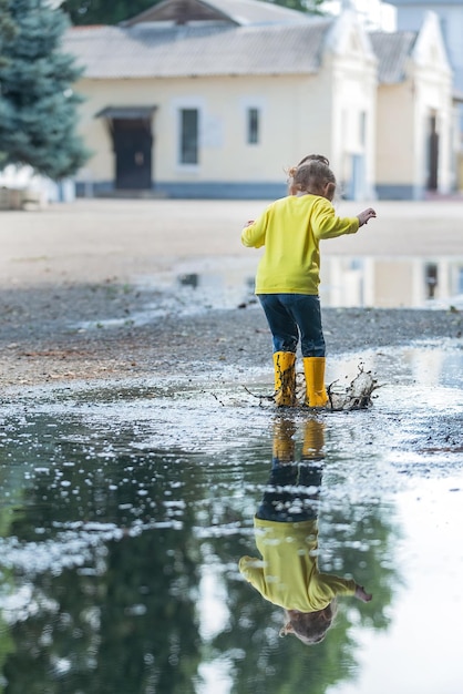 A little girl in yellow clothes and rubber boots runs merrily\
through the puddles