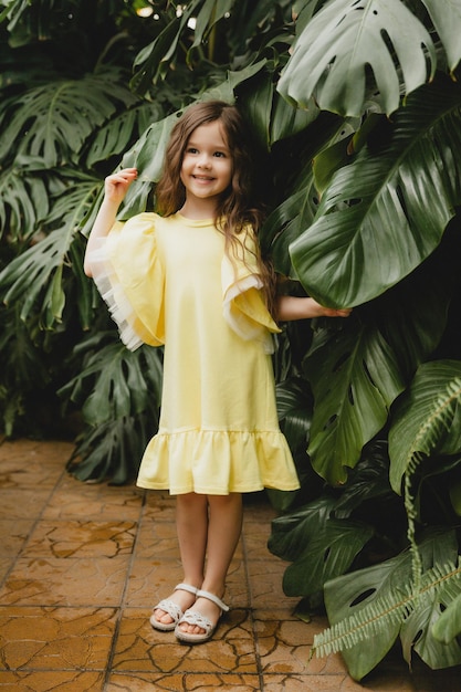 Little girl in a yellow dress in a botanical garden a child stands near the leaves of Monstera