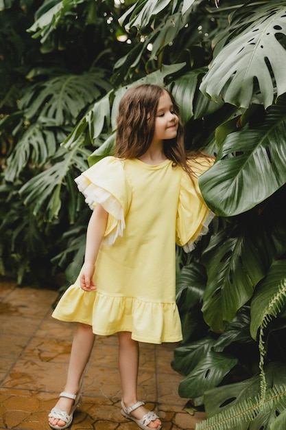 Little girl in a yellow dress in a botanical garden a child stands near the leaves of Monstera