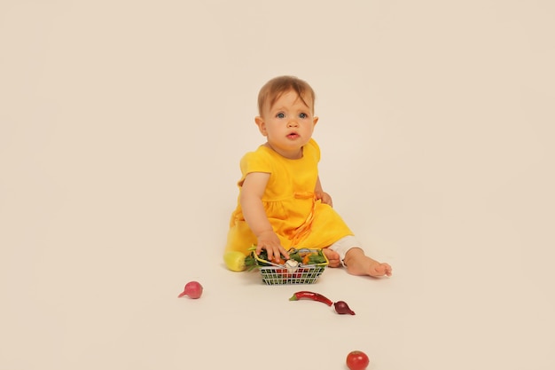 little girl in  yellow dress is sitting on a white background next to a small basket with vegetables