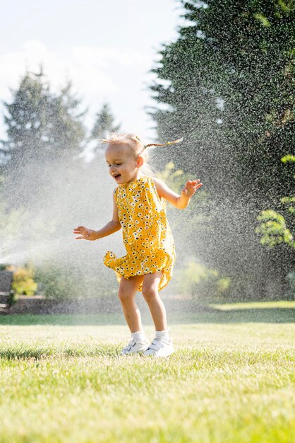 Photo a little girl in a yellow dress sprinkles a sprinkler.