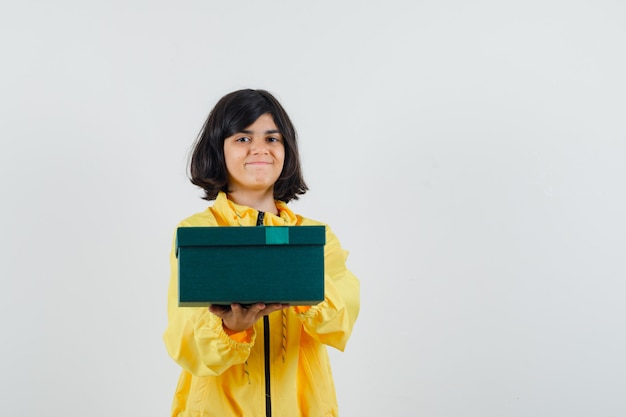 Little girl in yellow hoodie presenting gift box and looking cheerful , front view.