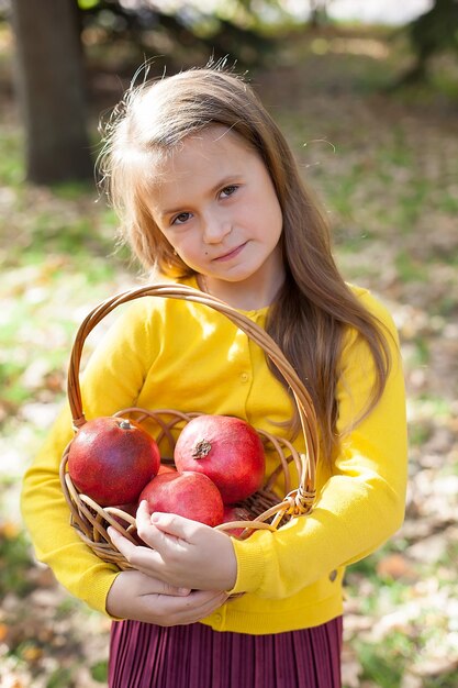 little girl in a yellow jacket and maroon skirt stands in the Park and holds ripe pomegranates.