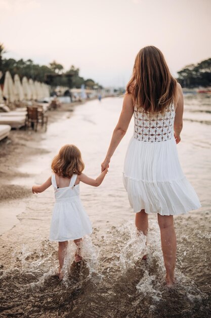 A little girl and a young mother in white dresses walk barefoot on the sand by the sea at sunset