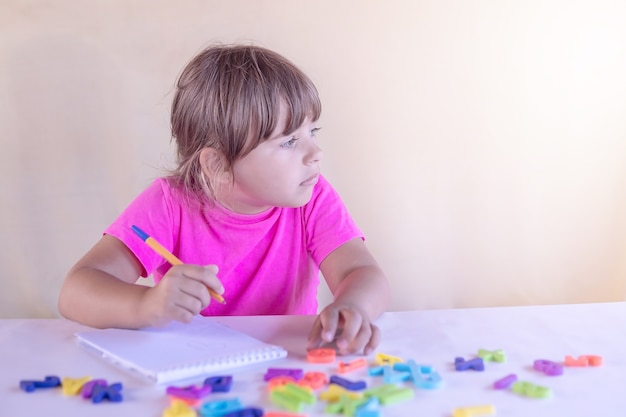 A little girlchild sits thoughtfully at a table with a notebook and letters and looks away