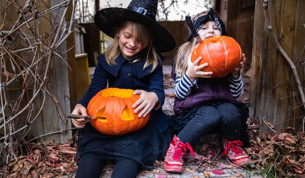 Маленькие девочки делают фонарь из больших тыкв для празднования ХэллоуинаКостюм ведьмы Шляпа Пальто Вырезать ножомВынуть мякоть с семечкамиЗадний двор для активного отдыхаДетская вечеринка