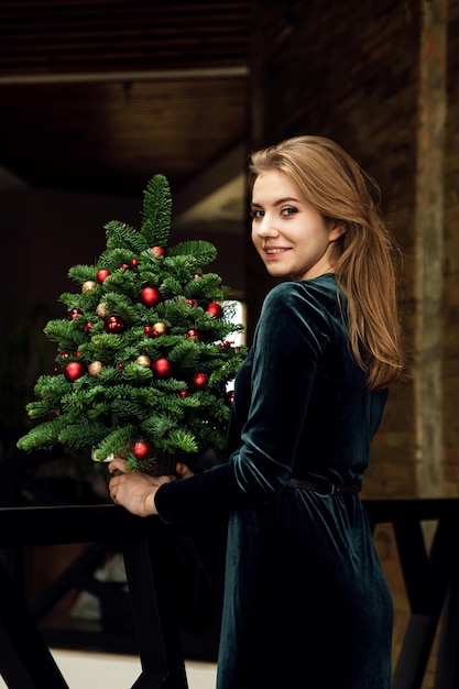 Маленькая зеленая рождественская елка украшена различными новогодними украшениями. рождественские каникулы. милая девушка в праздничном зеленом платье держит елку дома. домашняя рождественская атмосфера