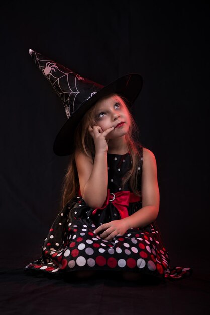 Little halloween witch in dark clothing sitting on black background and thinks.