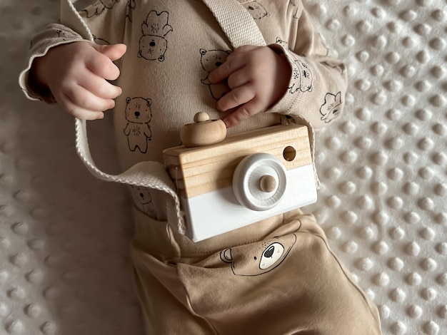 The little hands of a baby in a beige suit hold a wooden camera toy view from above cropped photo