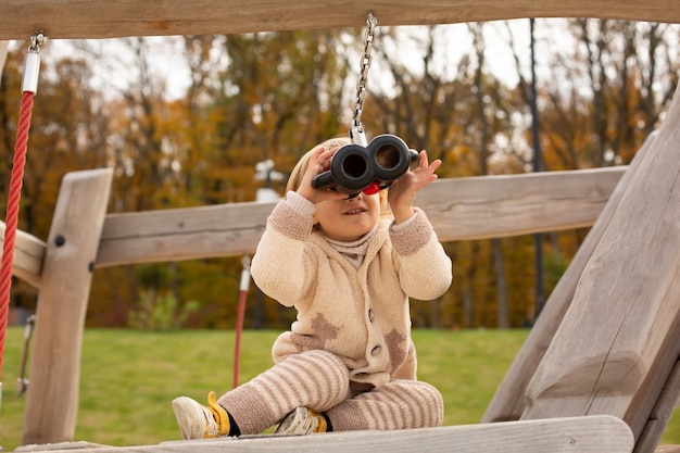 a little handsome boy in a light knitted warm suit looks through binoculars in the city