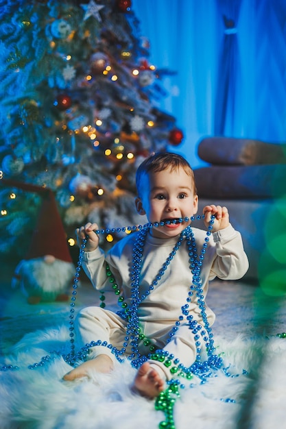 Little handsome oneyearold boy dressed in a beige suit on the background of a Christmas tree in a home environment Children's emotions before celebrating the New Year holiday