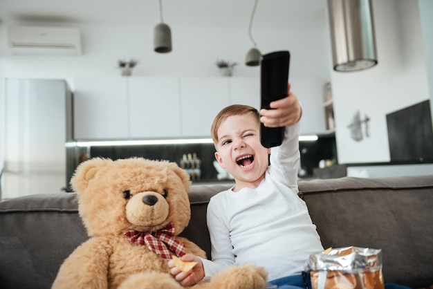 Little happy boy sitting on sofa with teddy bear at home and watching TV while eating chips. Holding remote control.