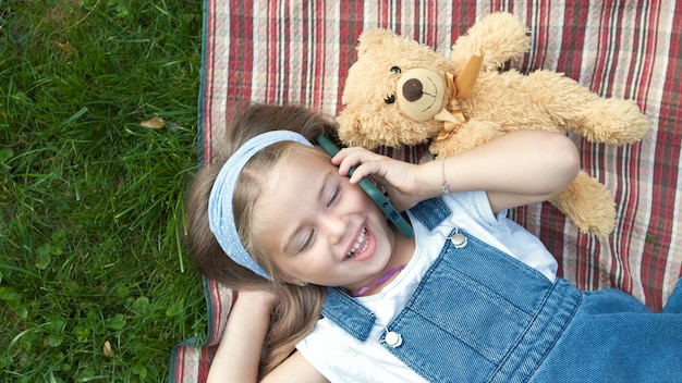 Little happy child girl laying on a blanket on green lawn in summer with her teddy bear talking on mobile phone.