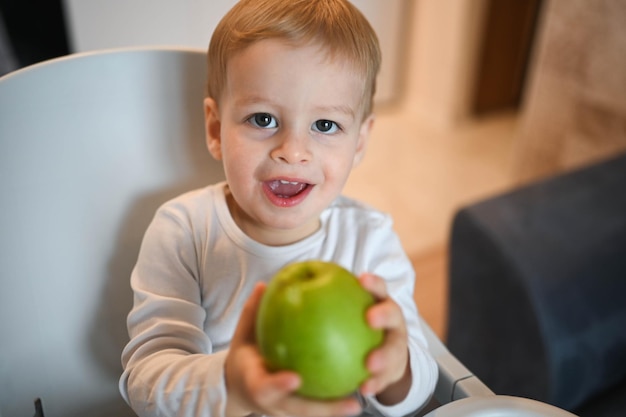 Little happy cute baby toddler boy blonde sitting on baby chair playing with apple baby facial