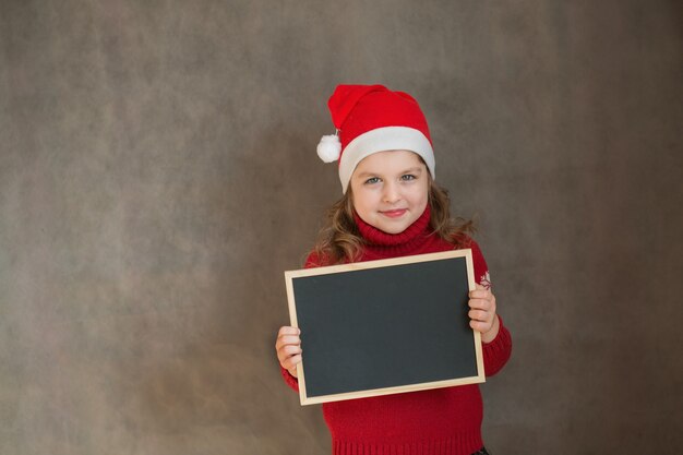 Little happy girl in Christmas jacket and santa hat with black desk