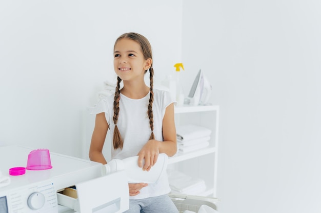 Little happy girl has two pigtails fills in washing machine\
with liquid detergent pours into tray of washer stands in laundry\
room with white walls helps mother with washing wears white t\
shirt