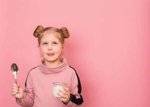 Little happy girl holding yoghurt and spoon on pink background