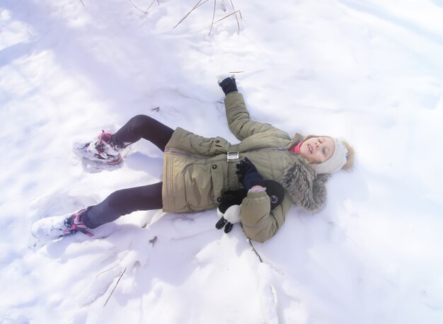 森の新雪の上に横たわっている小さな幸せな女の子。晴れた冬の日。