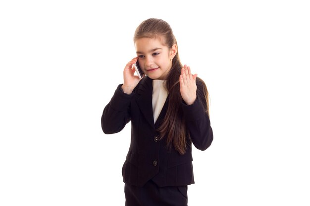 Little happy girl with long chestnut ponytail in black jacket and black skirt talking on the phone