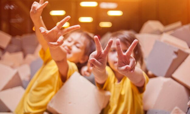 Bambine felici giocano e si divertono in una piscina a secco con cubetti di paralon in un centro di intrattenimento per bambini e mostrano le dita simbolo di pace e vittoria