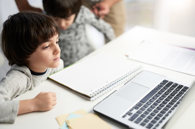 Ragazzino ispanico che sembra concentrato, ascolta l'insegnante durante la lezione online per bambini, seduto con suo fratello gemello al tavolo di casa. messa a fuoco selettiva