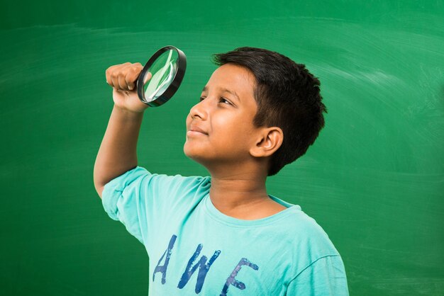 Little indian asian school boy holding magnifying glass standing isolated over green chalkboard background