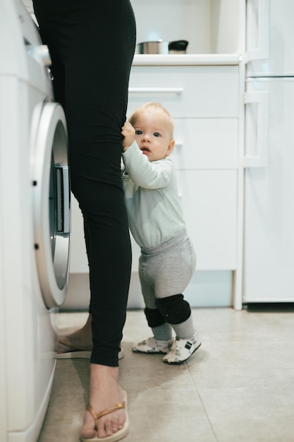 Little infant baby boy child hiding between mothers legs demanding her attention while she is multitasking trying to do some household chores in kitchen at home Mother on maternity leave