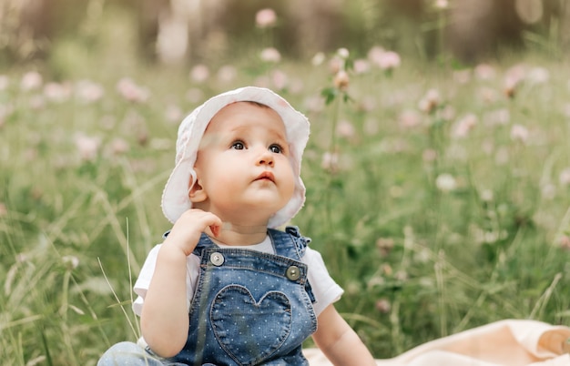 白い帽子をかぶった小さな幼児の女の子が花の間の公園に座っています