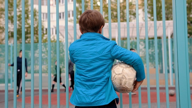 空気を抜いたボールを持って、他の子供たちがサッカーをしているのを見ている不安な少年