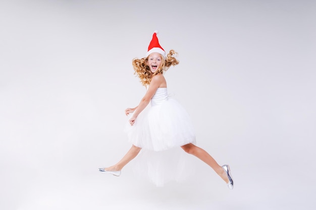 Little joyful girl in red Santa hat and white beautiful dress jumps from