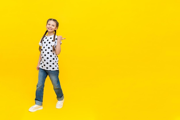 A little joyful girl with beautiful long pigtails in jeans on a yellow background